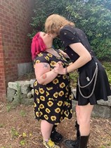 Photo of Kaelana kissing her partner Penny.  Kaelana wears a black sunflower dress and Penny is leaning over, wearing a black dress with a thin metal chain.