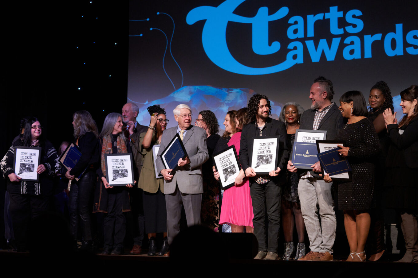 Awardees standing on a stage holding their awards and looking at each other with excitement.