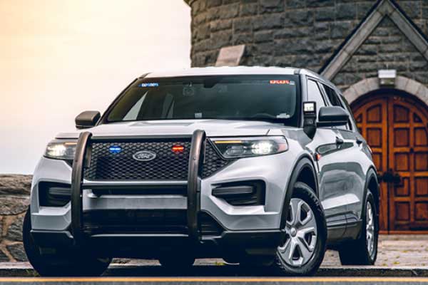 A gray Ford Explorer Police Interceptor Utility vehicle is parked in front of a stone building with a large wooden door. The vehicle has blue and red lights activated on its roof and grill. A cloudy sky provides the backdrop.